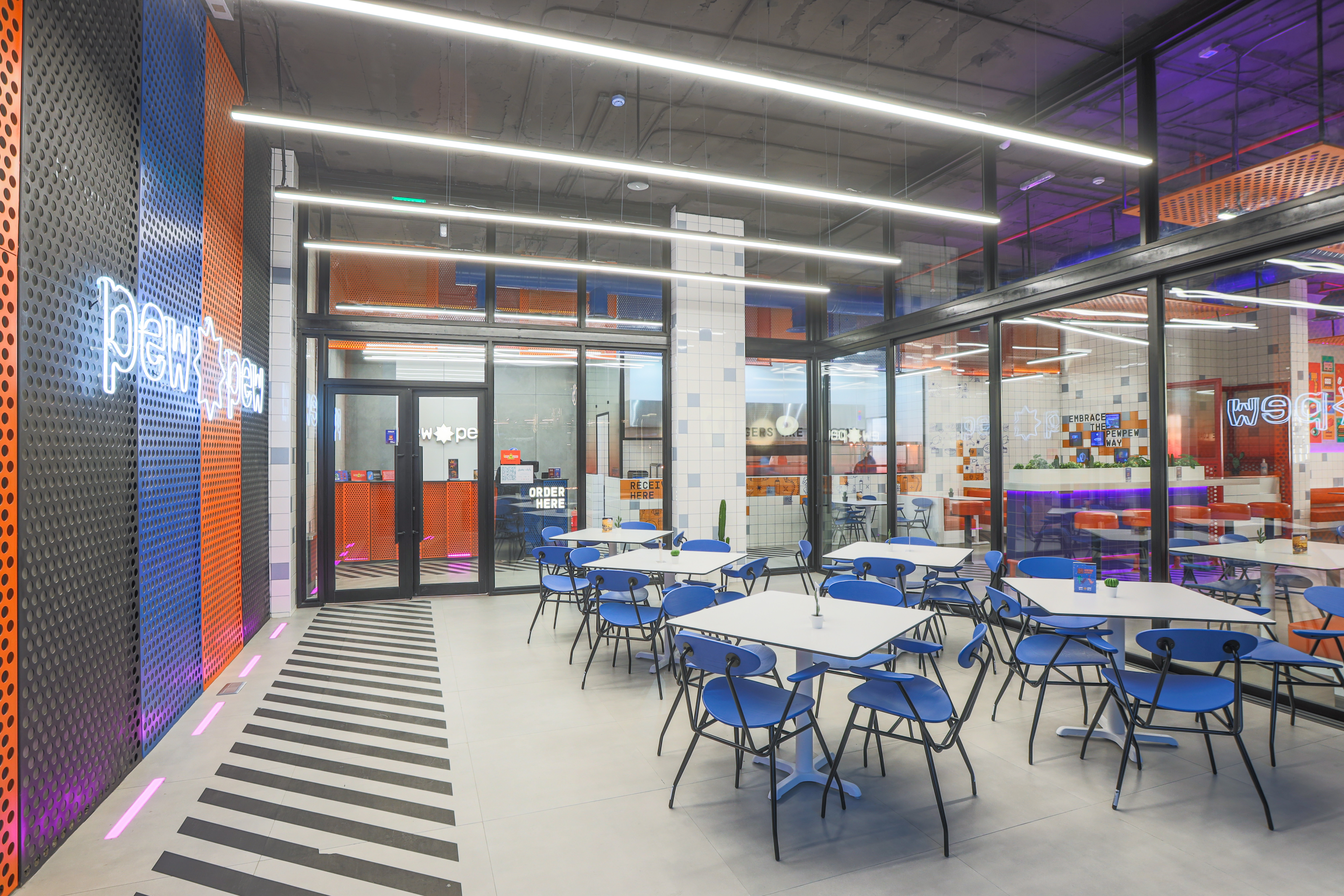 Pew Pew — dining hall with orange banquettes, blue chairs, and industrial ceiling